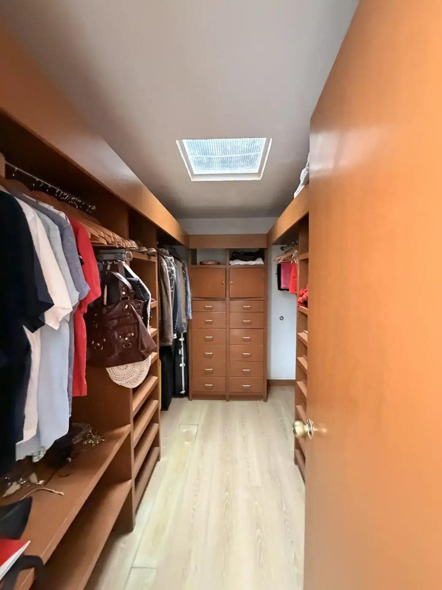 Amplio vestidor con estantes de madera, ropa organizada, cajoneras y un tragaluz en el techo, piso claro.