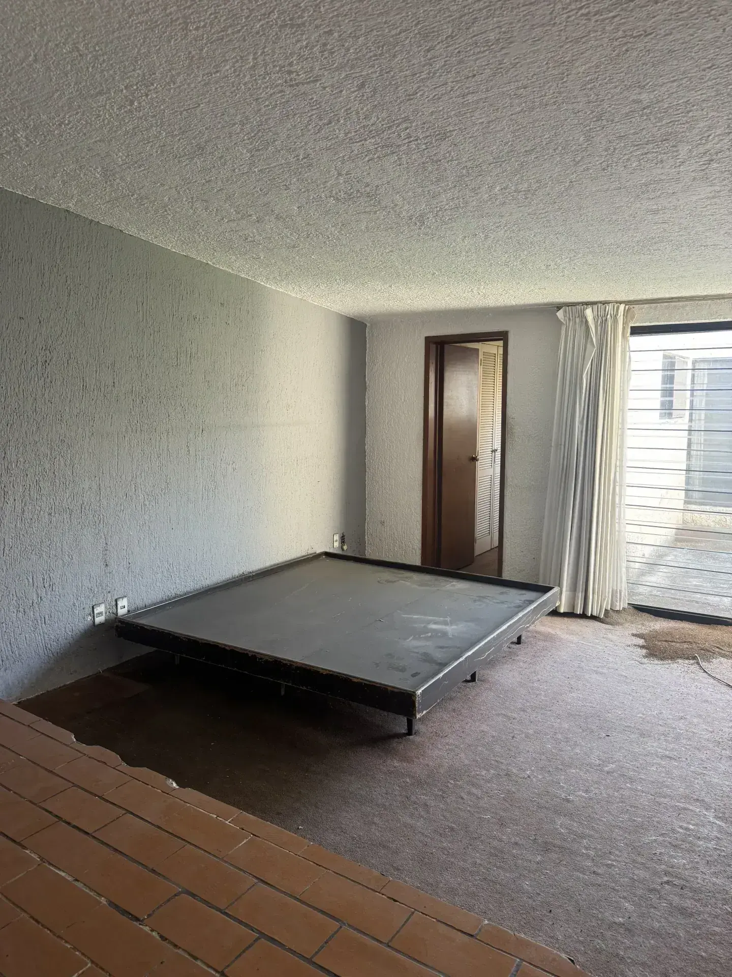 Habitación vacía con paredes grises texturizadas, alfombra sucia, base de cama y ventana, ideal para demoliciones en Zapopan.